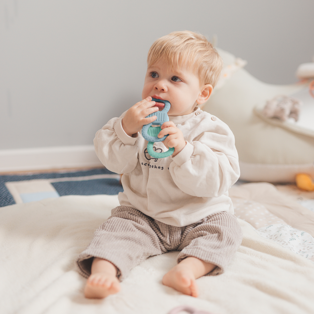 Glücklich lachendes Baby, das einen mintgrünen Beißring festhält und zum Mund führt.