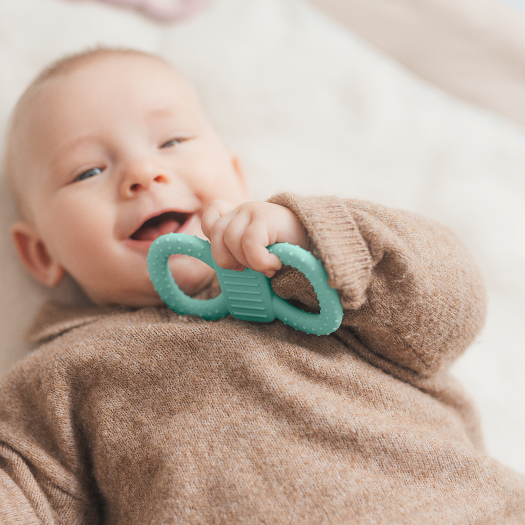 Ein fröhlich lächelndes Baby in einem braunen Strickpullover liegt auf einer weichen Decke und hält einen mintfarbenen, strukturierten Beißring in der Hand.