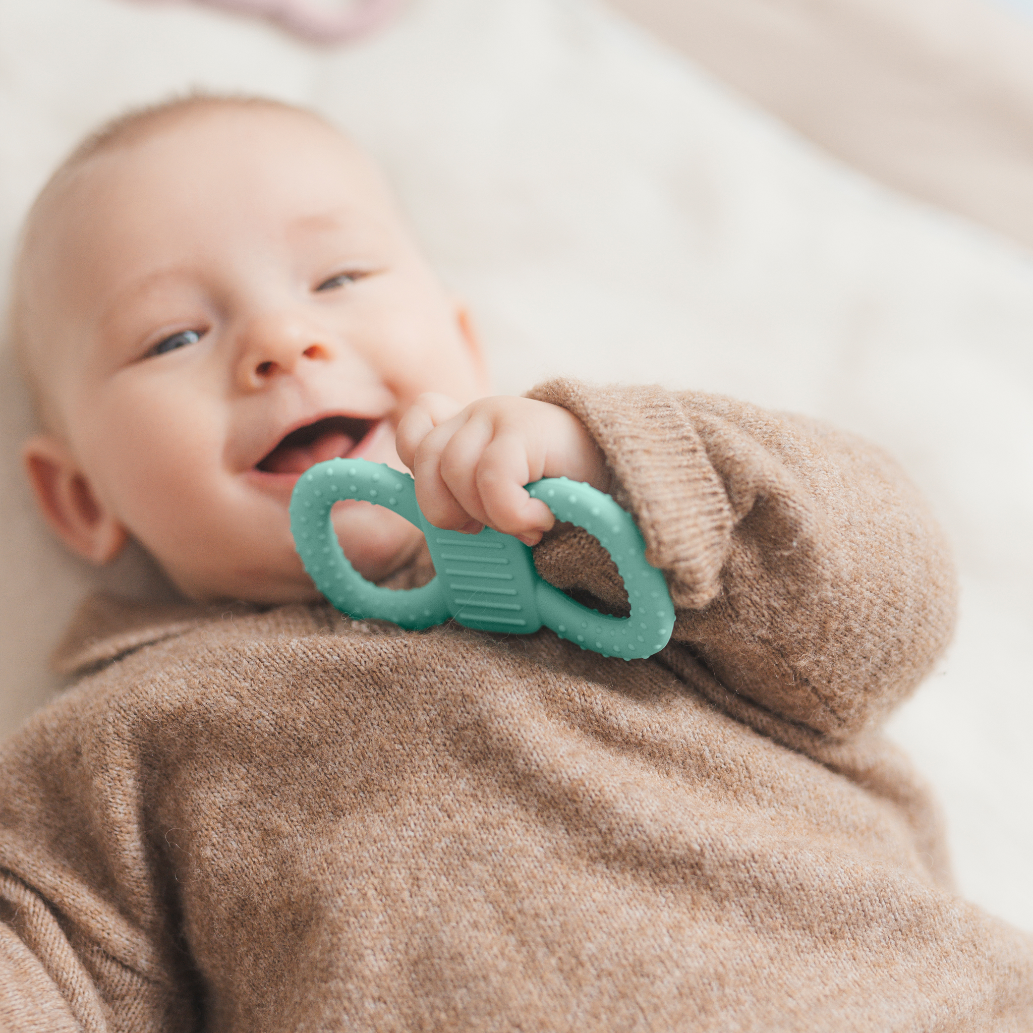 Ein fröhlich lächelndes Baby in einem braunen Strickpullover liegt auf einer weichen Decke und hält einen mintfarbenen, strukturierten Beißring in der Hand.