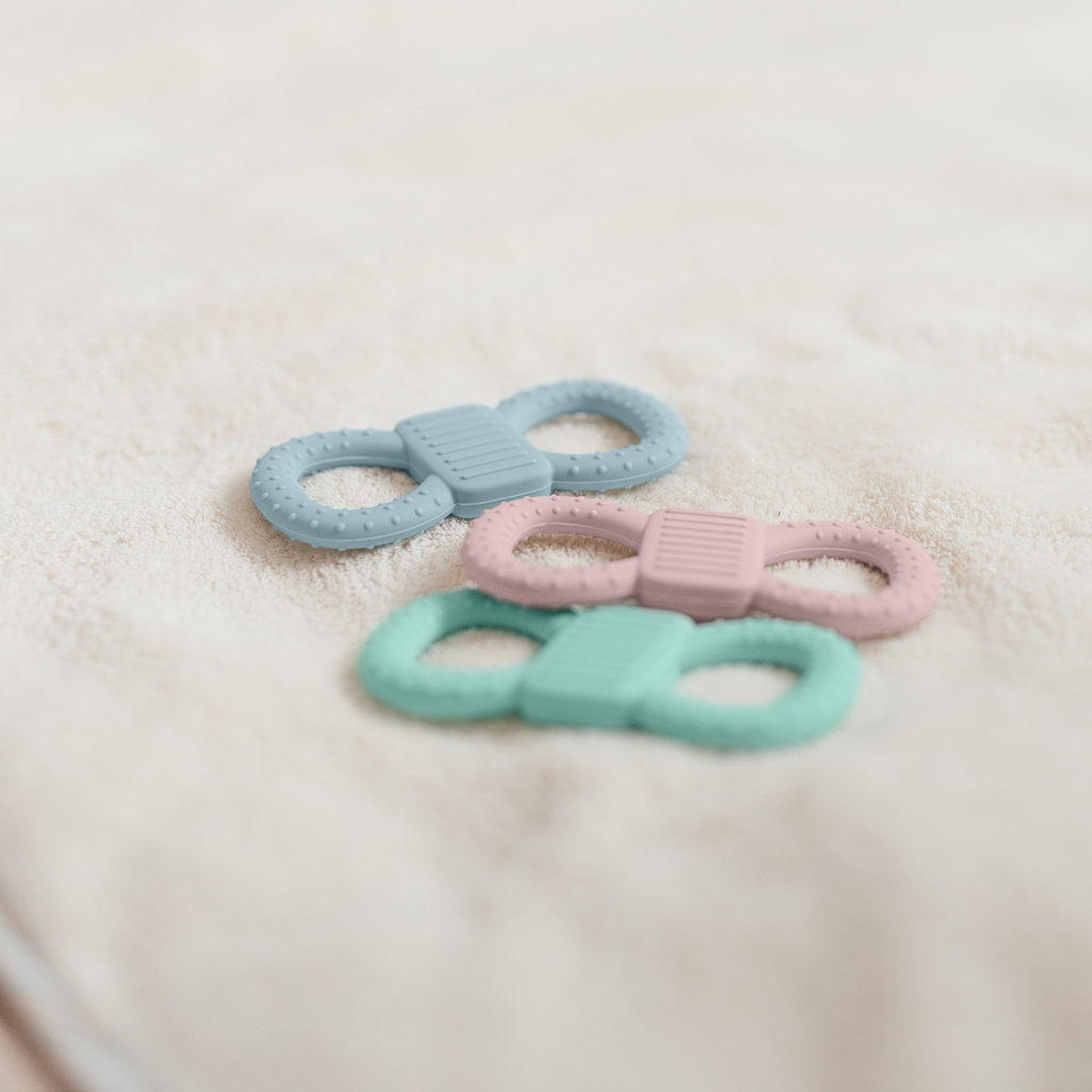 Drei Beißringe in den Farben Hellblau, Rosa und Mint, die leicht versetzt hintereinander auf einer weichen, beigen Frottee-Decke liegen.