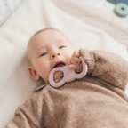 Ein Baby in einem braunen Wollpullover liegt auf einer hellen Decke und beißt auf einen rosafarbenen, strukturierten Beißring.